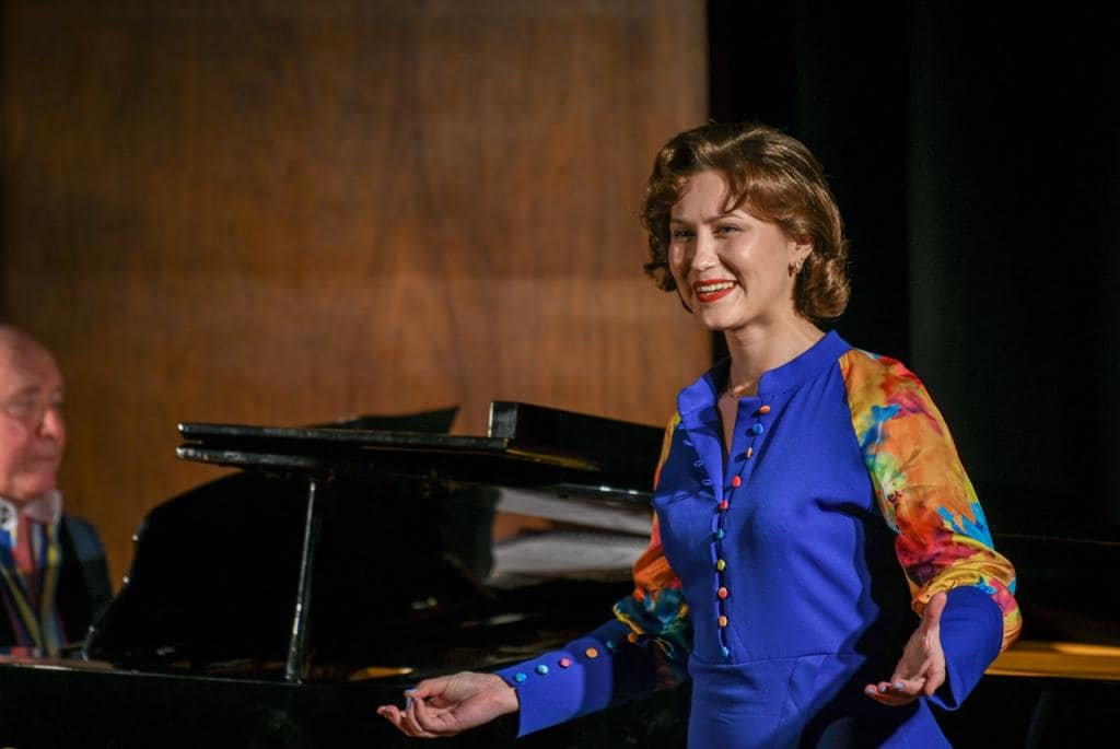 A woman with short curly hair wearing a colorful blouse stands next to a piano, smiling, with a man in the background.
