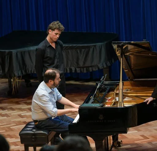 A young man standing and a man sitting at a grand piano during a performance or rehearsal in a music hall.