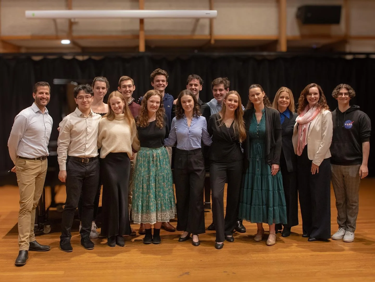 Group of people, both men and women, standing on a wooden floor in front of a black curtain, posing for a photo.