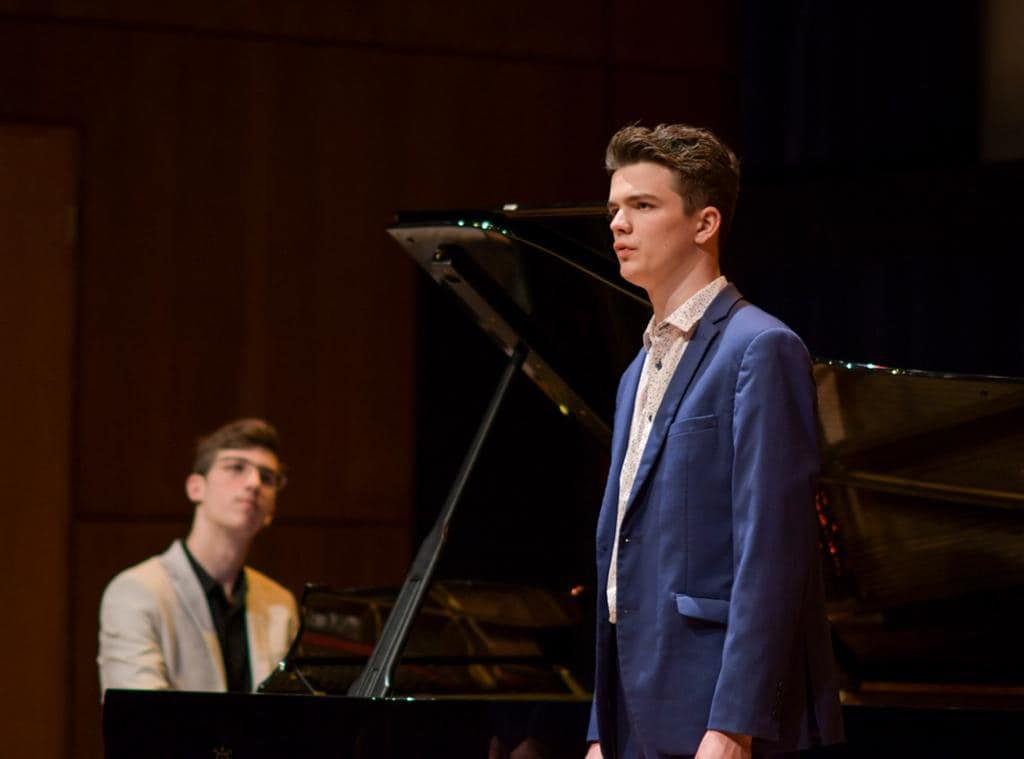Two young men in formal attire, one in a beige blazer and the other in a blue blazer, performing on a stage with a grand piano. The man in the blue blazer is standing and appears focused, while the other is seated at the piano.
