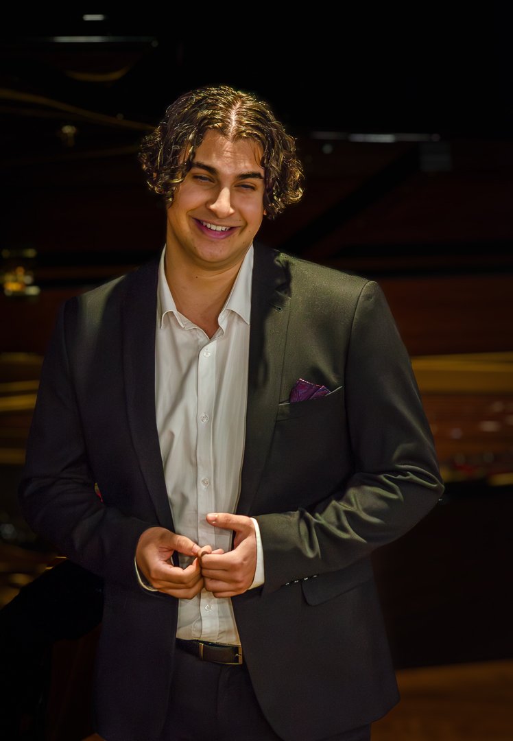 Young man with curly hair in a suit, smiling at the camera, standing indoors with a dark wooden background.