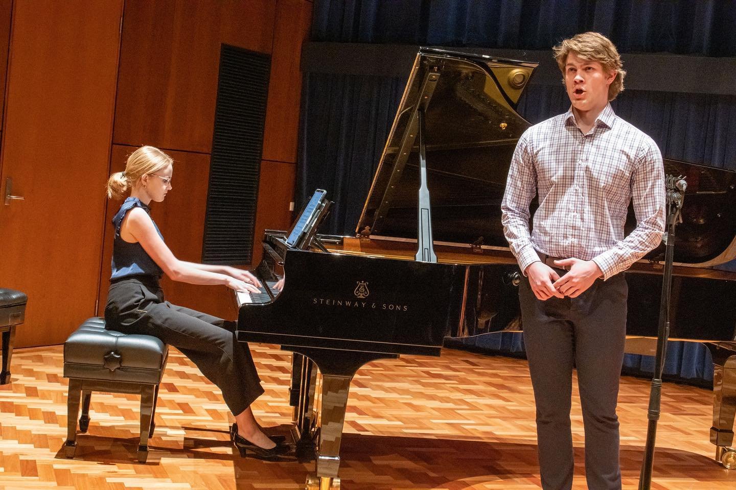 A woman playing a grand piano with a man singing next to her, in a concert hall setting.