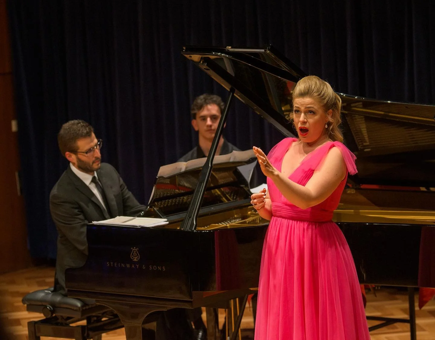 A female singer in a bright pink gown performs in front of a grand piano, while two male pianists accompany her on stage.