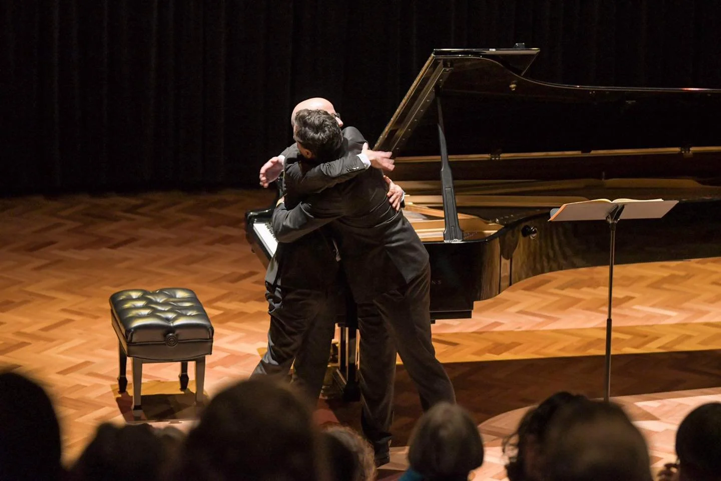 Two men are hugging on stage in front of a grand piano, with an audience in the foreground.