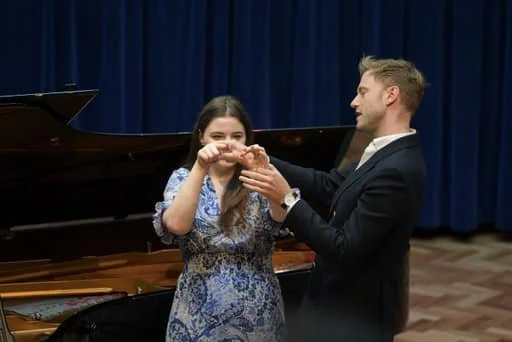 A man handing a ring to a young woman in front of a grand piano and blue curtain