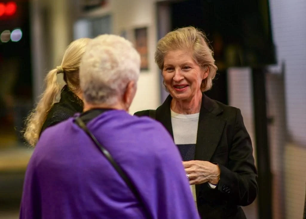 Three women engaged in conversation indoors, one smiling and wearing a black blazer, another with gray hair in a purple top, and a blonde woman with a ponytail.