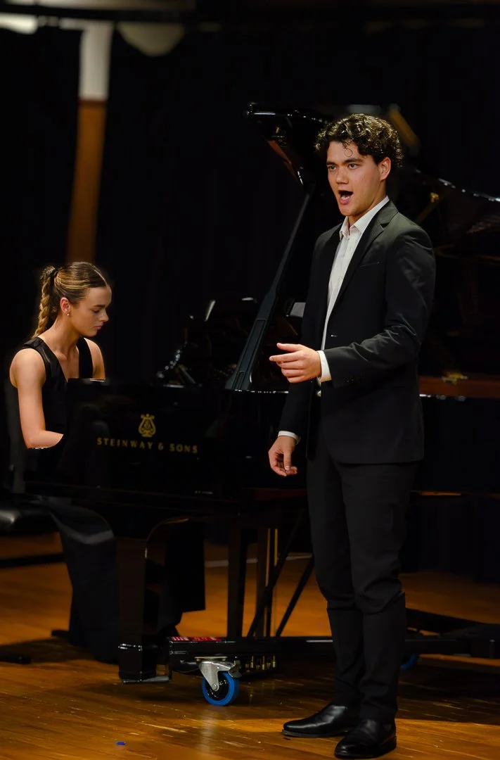 A male singer in a black tuxedo performs in front of a female pianist at a concert, with a grand piano on a wooden stage.