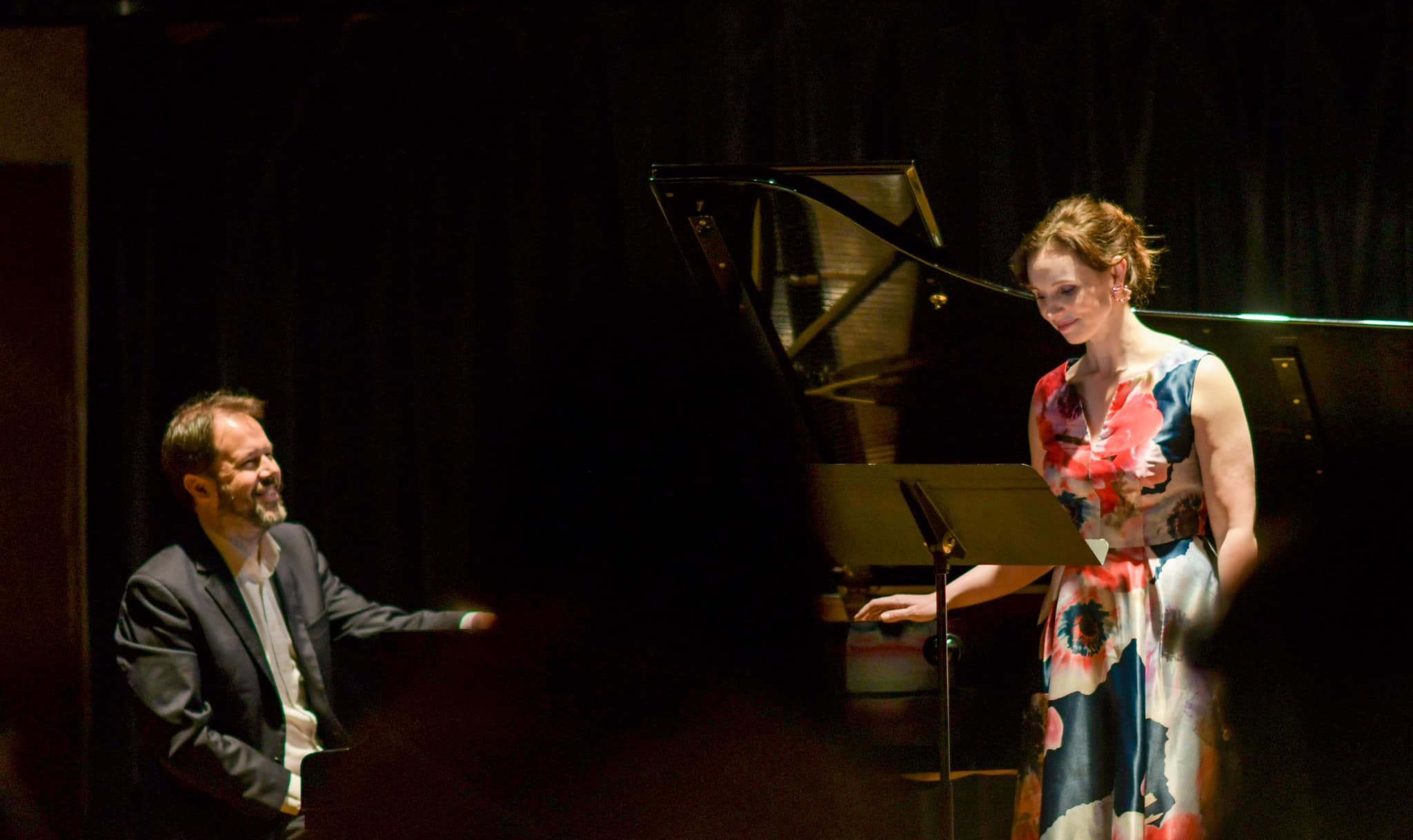 A man playing a grand piano and a woman standing next to the piano, holding a sheet of music, on a dark stage.