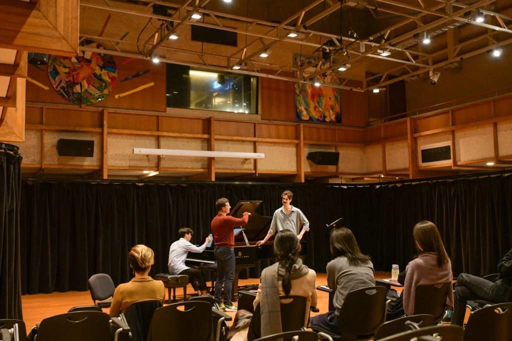 A group of people in a music rehearsal room with black curtains, a grand piano, and three men practicing or performing while the audience watches.