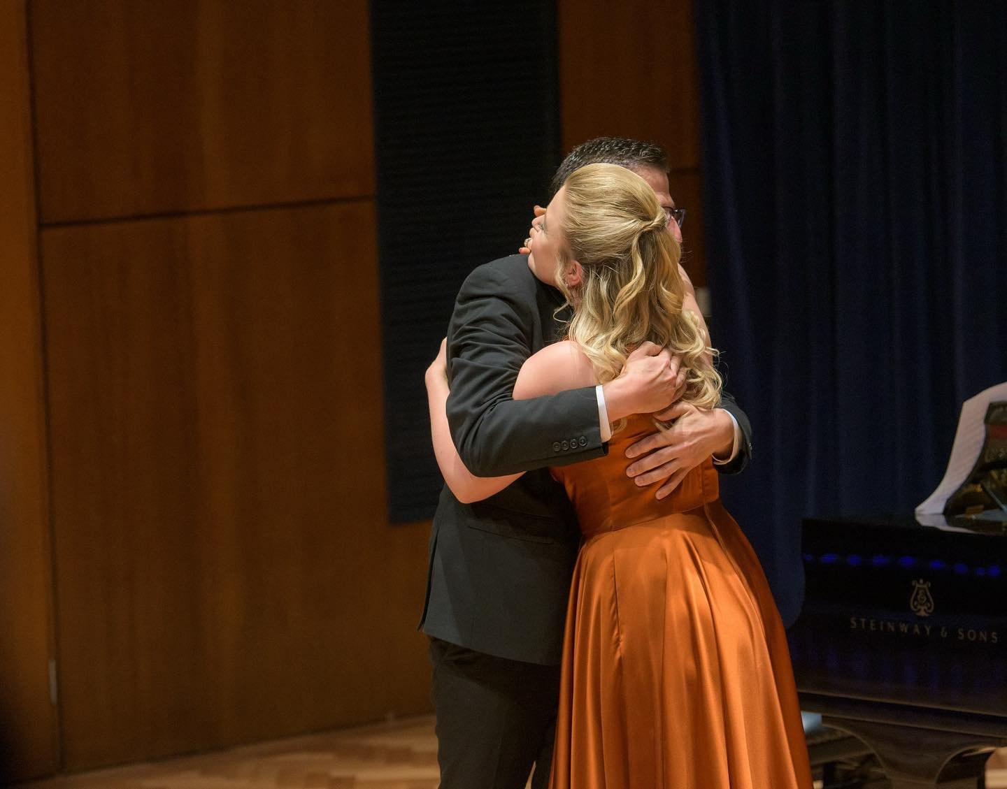 A couple hugging closely, possibly on stage, with a piano in the background.