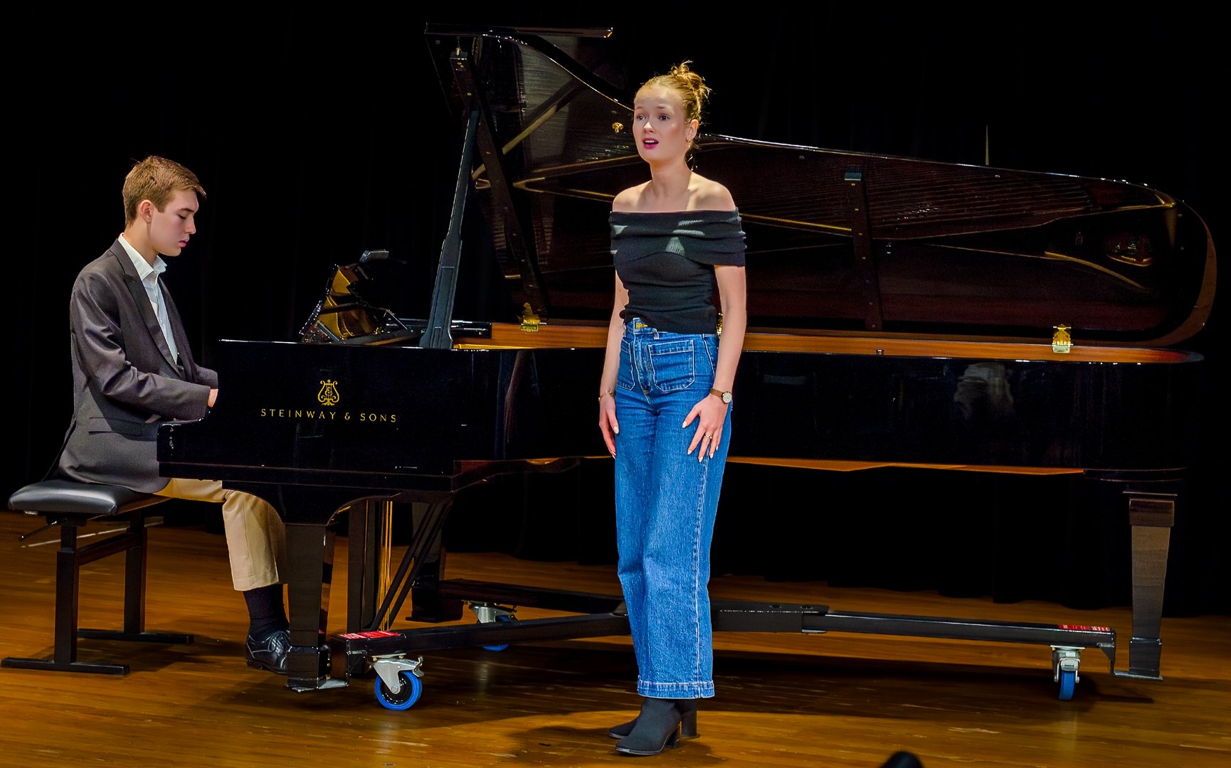 A woman singing in front of a grand piano with a man playing the piano on stage with a black background.