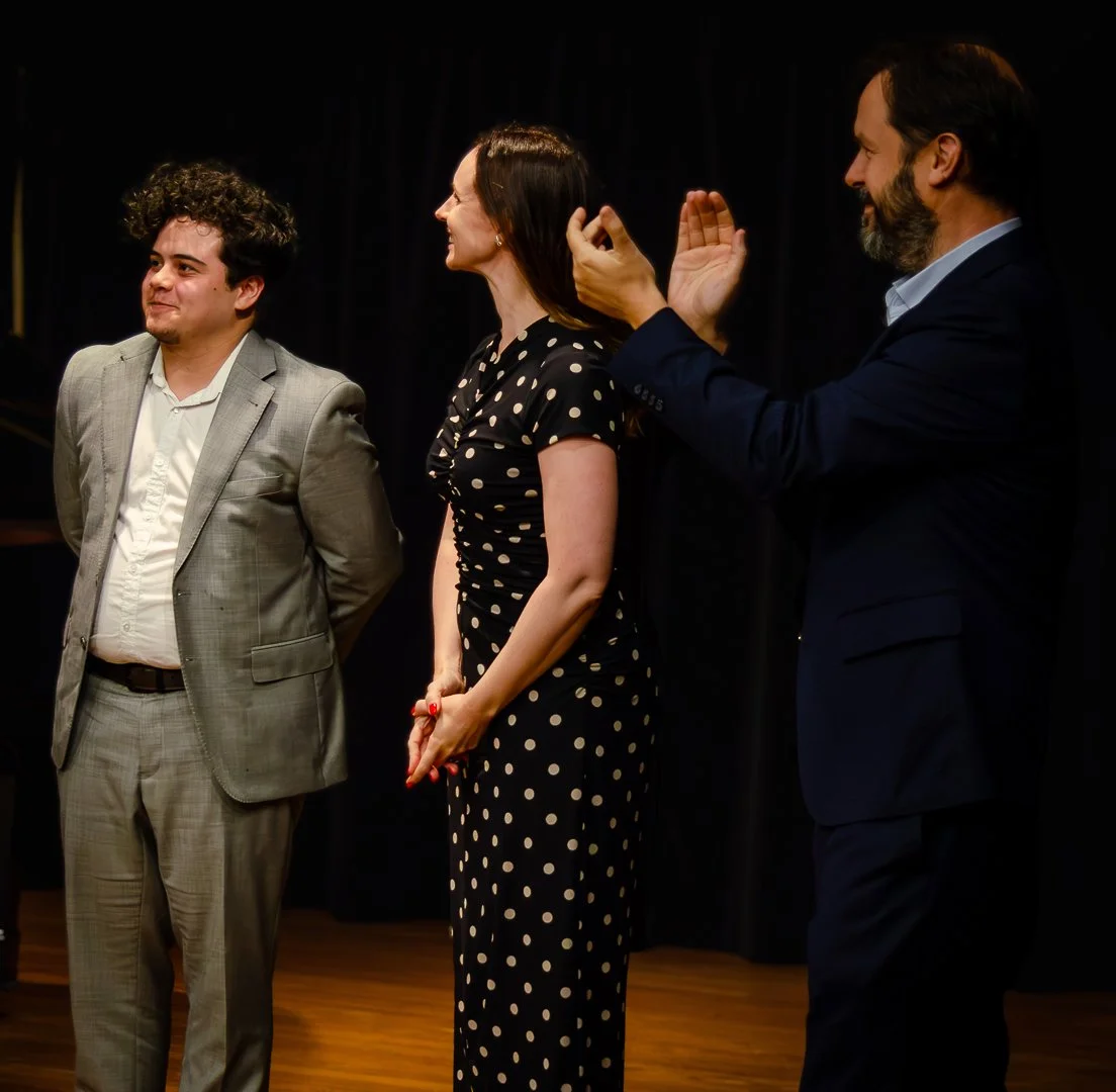 Three people on stage, two men and one woman, dressed in formal attire, with the woman receiving applause.