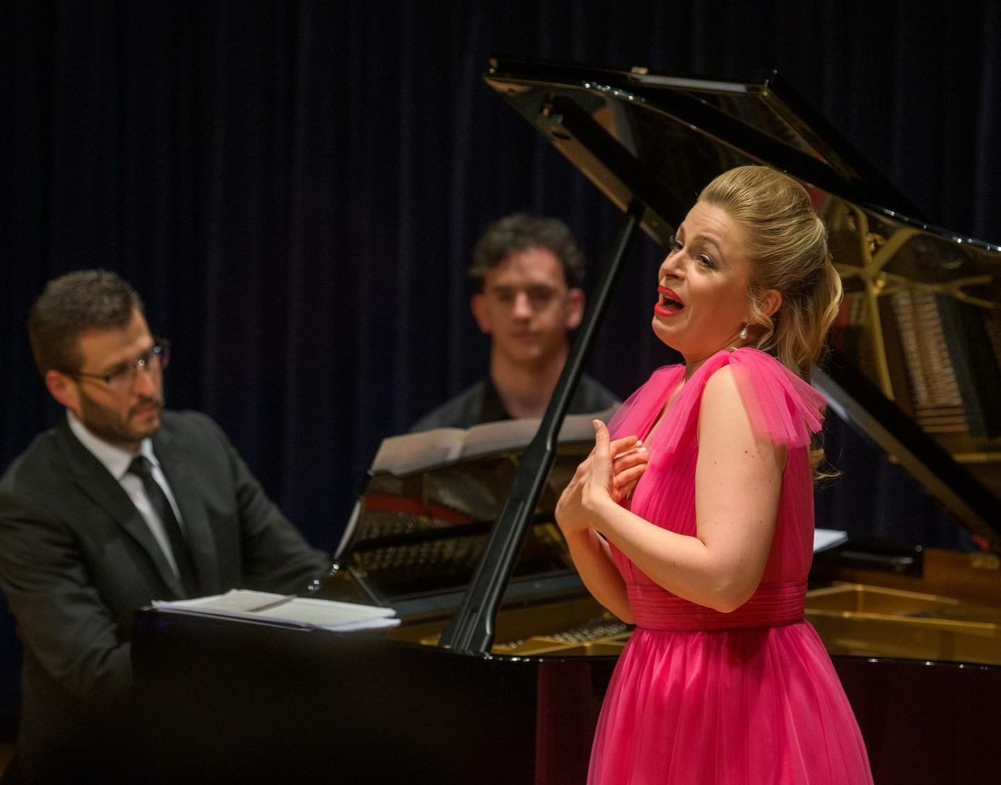 A woman in a bright pink dress singing passionately with her hand on her chest, accompanied by a pianist and a man observing, on a stage with dark curtains.