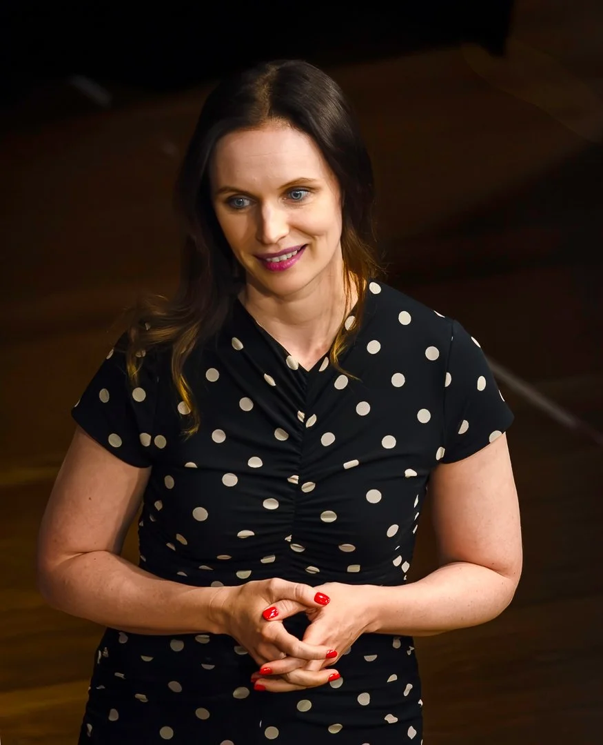 A woman with long dark brown hair, blue eyes, and pink lipstick smiling, wearing a black dress with white polka dots and red nail polish, standing indoors.