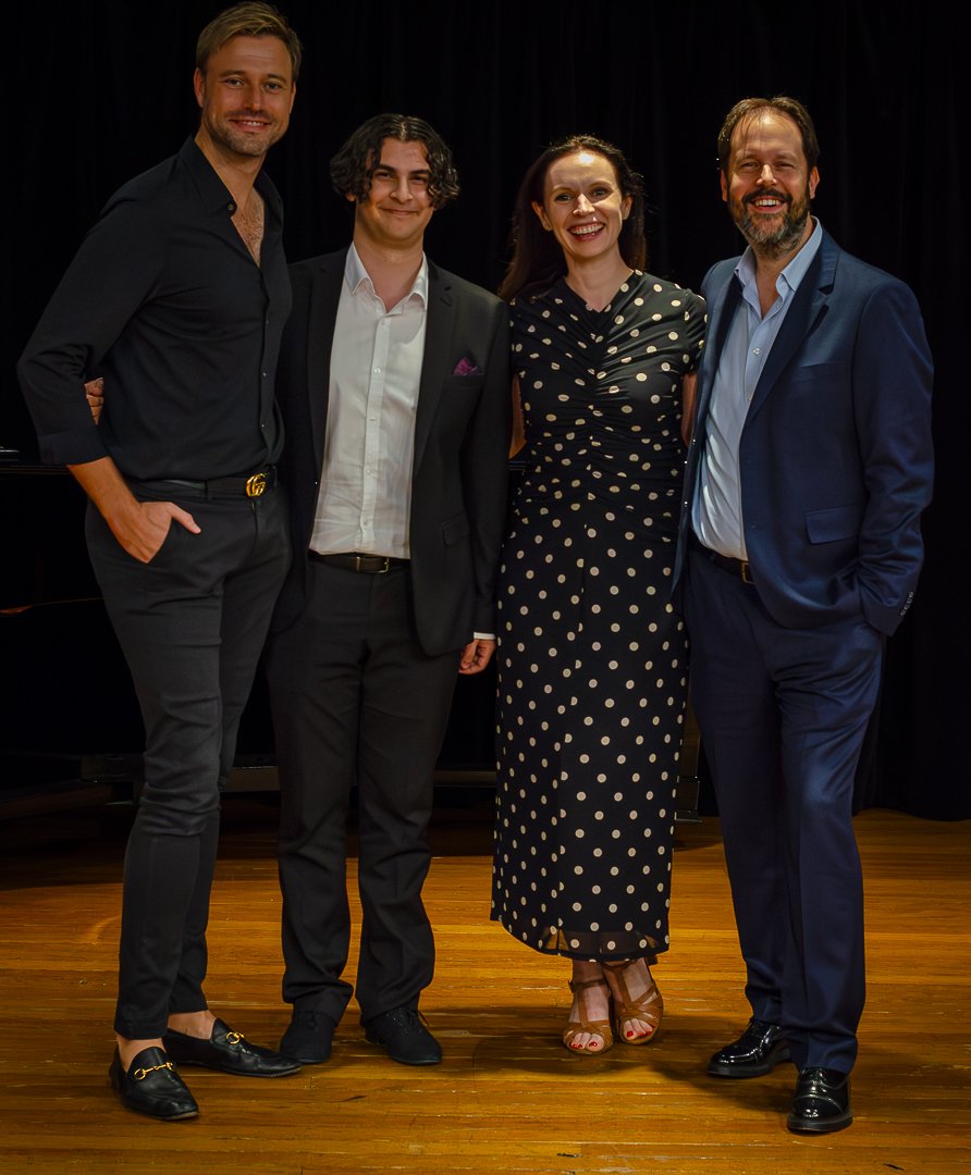 Four people standing together on a wooden stage, smiling for the camera, against a black curtain backdrop. The group includes three men and one woman, dressed in formal and semi-formal attire.