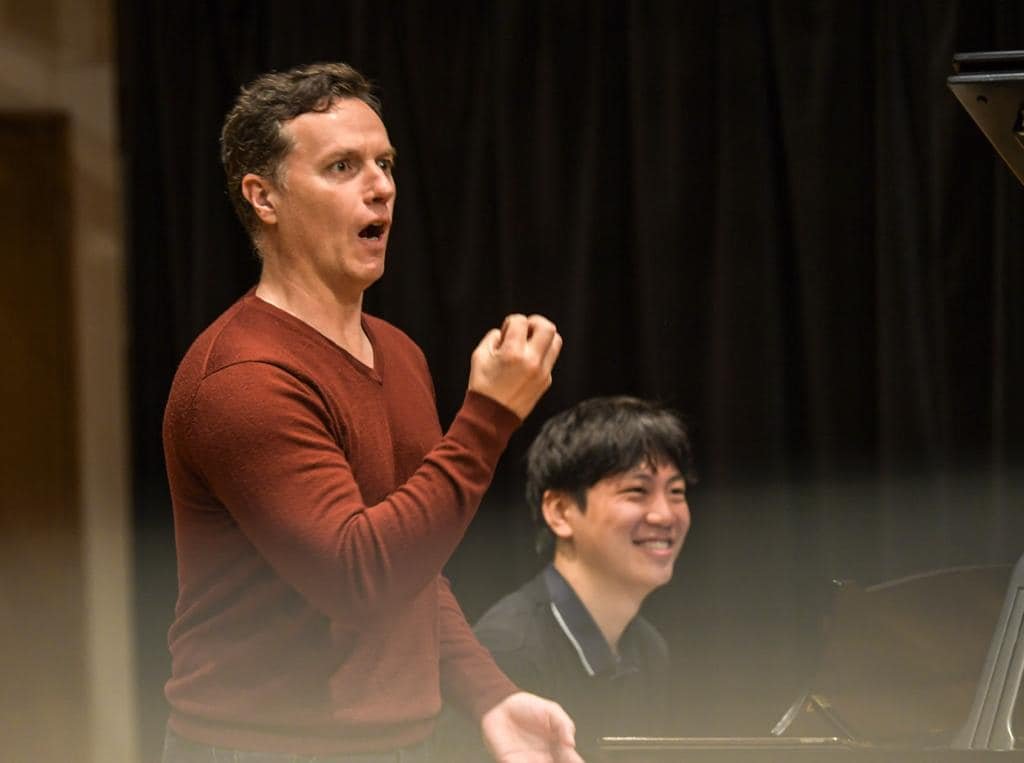 Two men in a music studio, one standing and making a gesture with his hand, the other sitting at a piano and smiling.