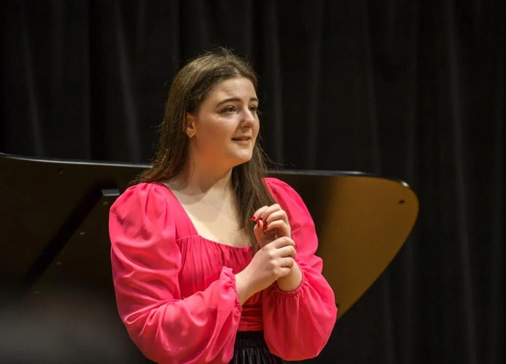 Young woman with long brown hair, wearing a pink dress, standing in front of a black curtain, possibly on stage or in a theater, with hands clasped.