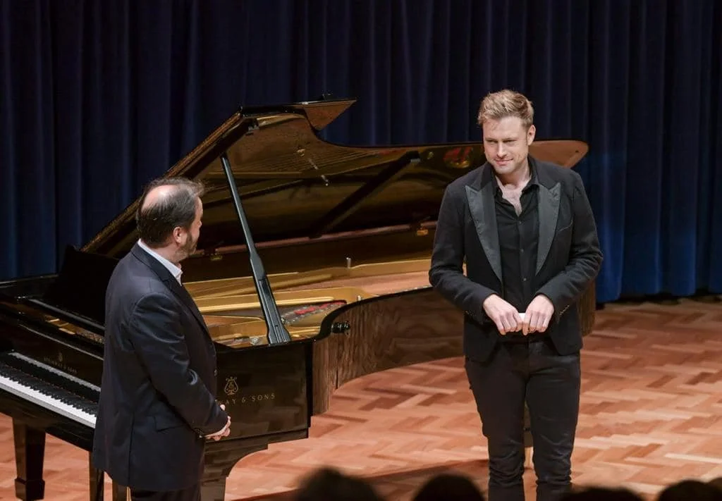 A man in a black jacket standing on a stage with a grand piano behind him, facing a man in a suit, in front of dark blue curtains.