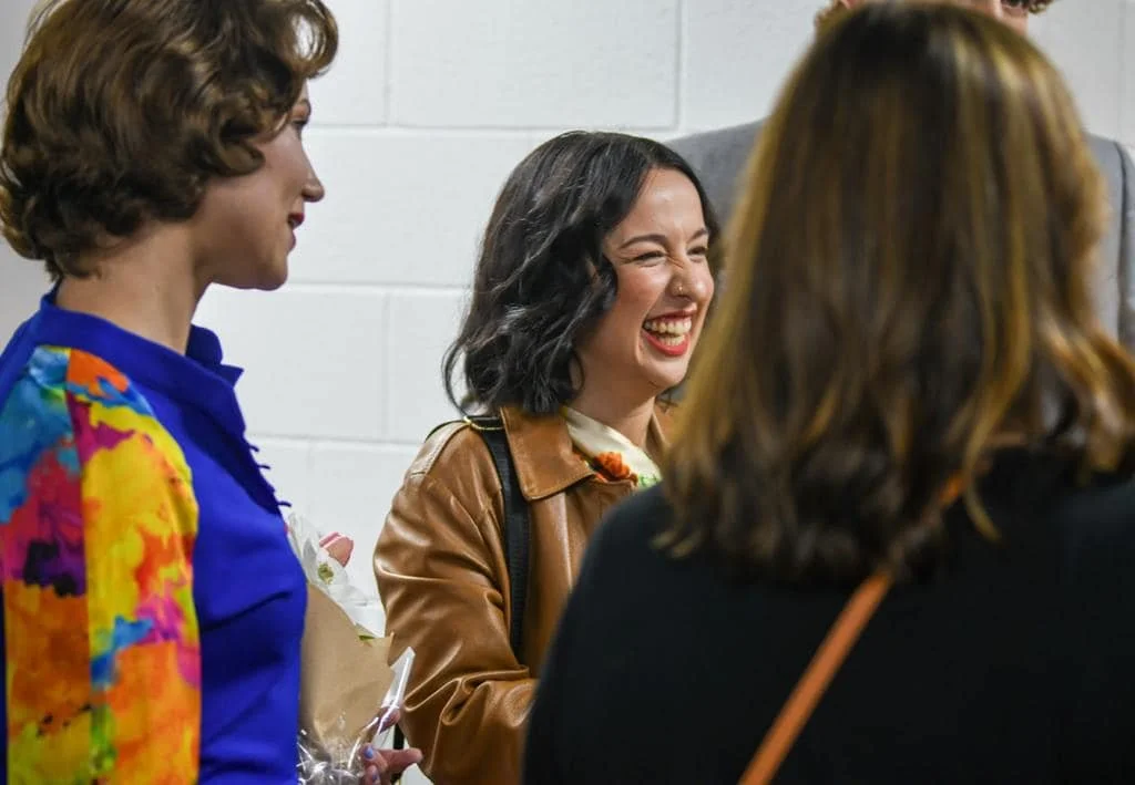 Group of women laughing and talking in a casual setting, one woman wearing a colorful jacket, another with a brown leather jacket.