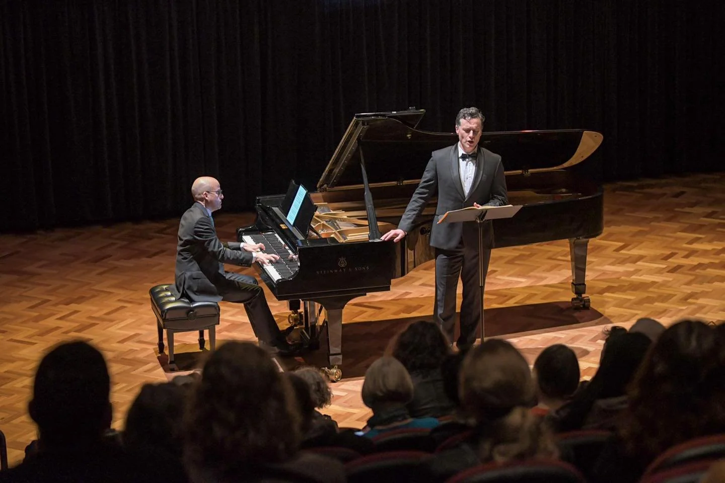 A pianist and a singer perform on a concert stage with a grand piano, in front of an audience.