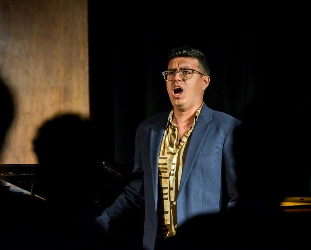 A man singing passionately at a piano in a dimly lit performance setting.