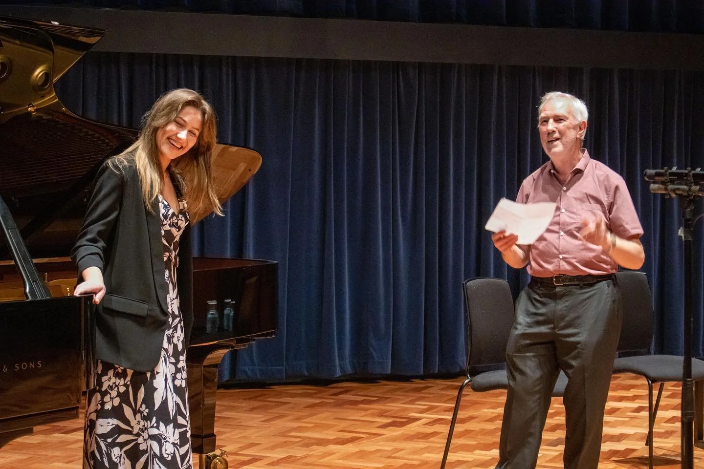 A young woman and an older man sharing a joyful moment on a stage with a grand piano and blue curtains in the background.