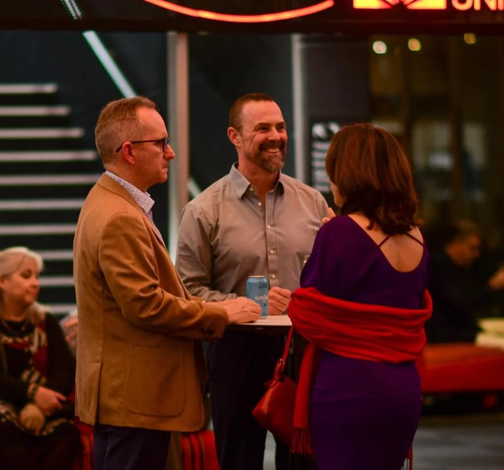Three people are standing and talking in a social setting, with a woman facing away from the camera and two men facing her, one of whom is smiling. They are indoors with blurred background and stairs visible behind them.
