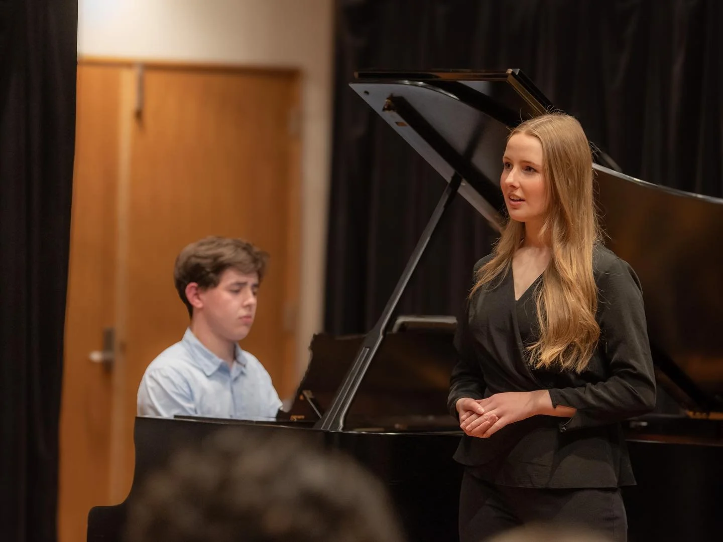 Young woman singing on stage with a grand piano and a young man playing the piano in the background.