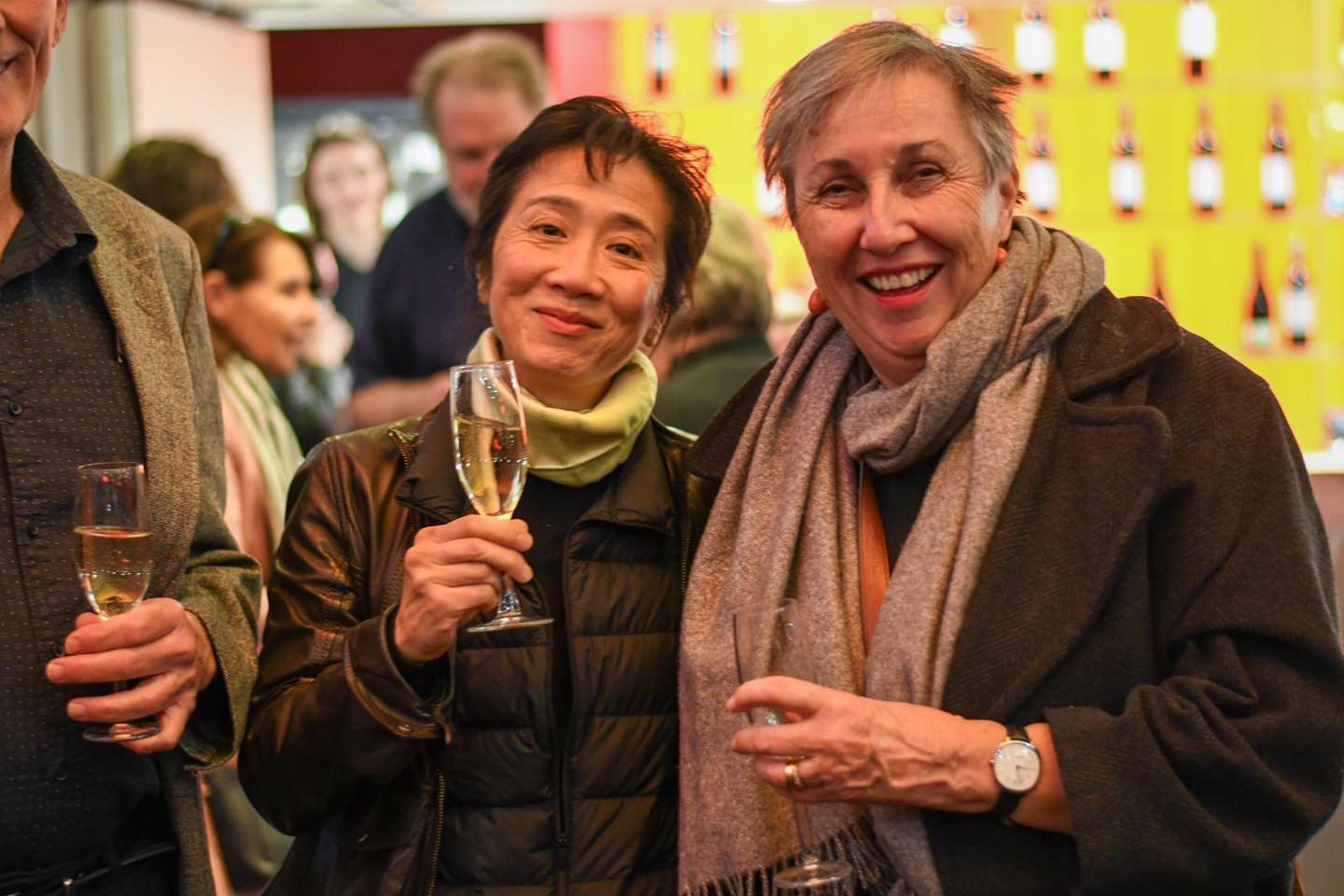 Two women smiling and holding glasses of champagne at a social gathering, with a colorful background and other people in the background.