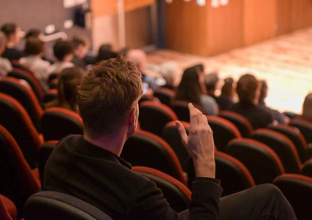 Person raising hand in audience during a presentation in a theater with a blurred stage in the background.
