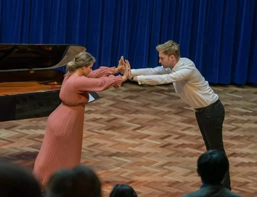 A woman in a pink dress and a man in a white shirt and black pants are performing a dance move, facing each other with their arms extended and hands pressed together, on a wooden floor with a blue curtain backdrop.