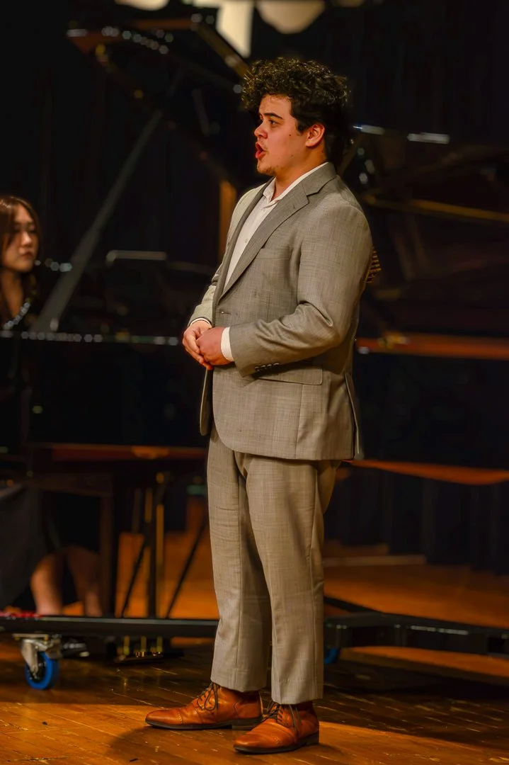 A young man in a light gray suit and brown shoes standing on stage in front of a grand piano, possibly performing or giving a speech.