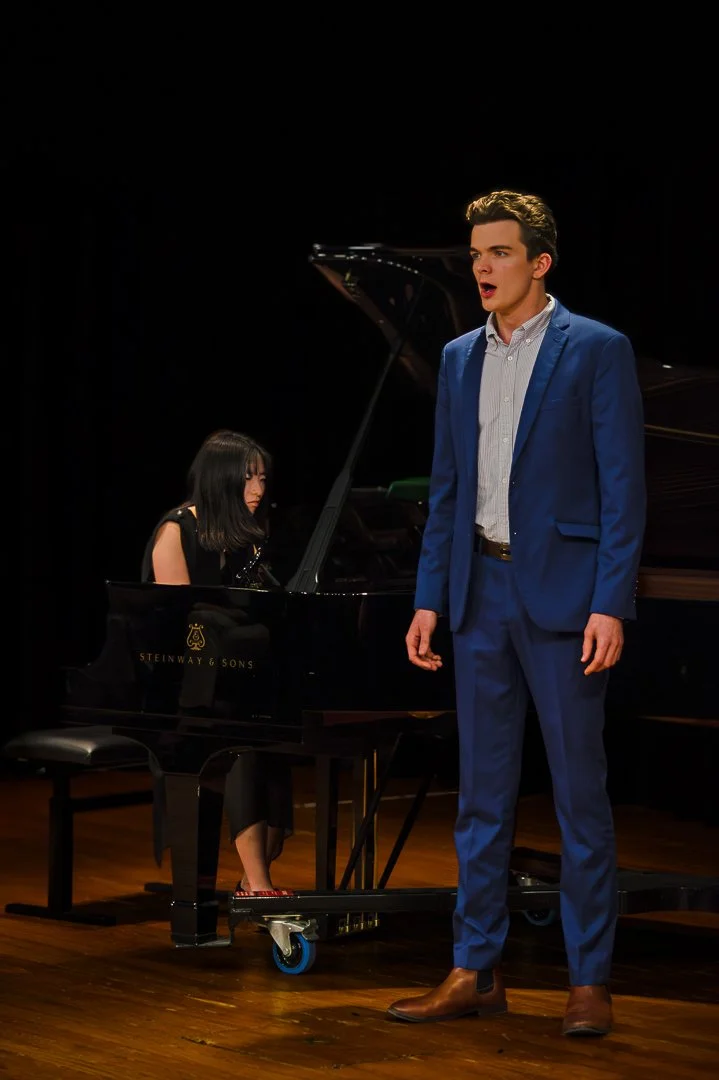 A man in a blue suit singing while a woman plays a grand piano on stage.