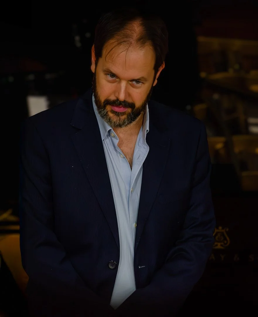 A man with a beard and dark hair wearing a dark blazer and light blue shirt, looking directly at the camera with a slight smile.