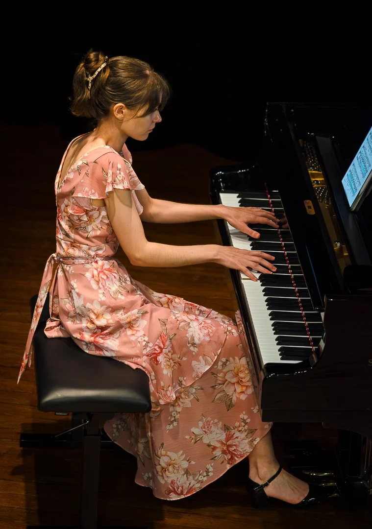 A woman in a pink floral dress playing a grand piano on a stage.