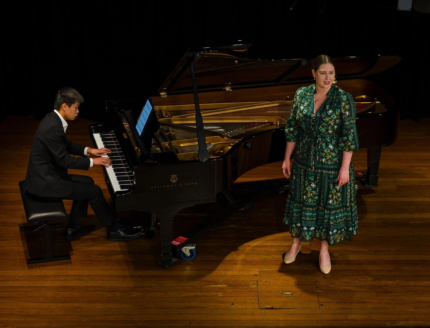 A woman in a long floral dress singing on stage while a man plays a grand piano