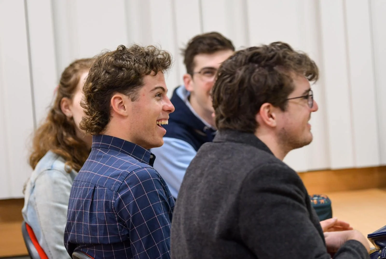 Group of four young adults sitting and smiling in a classroom or lecture hall with wood-paneled walls.