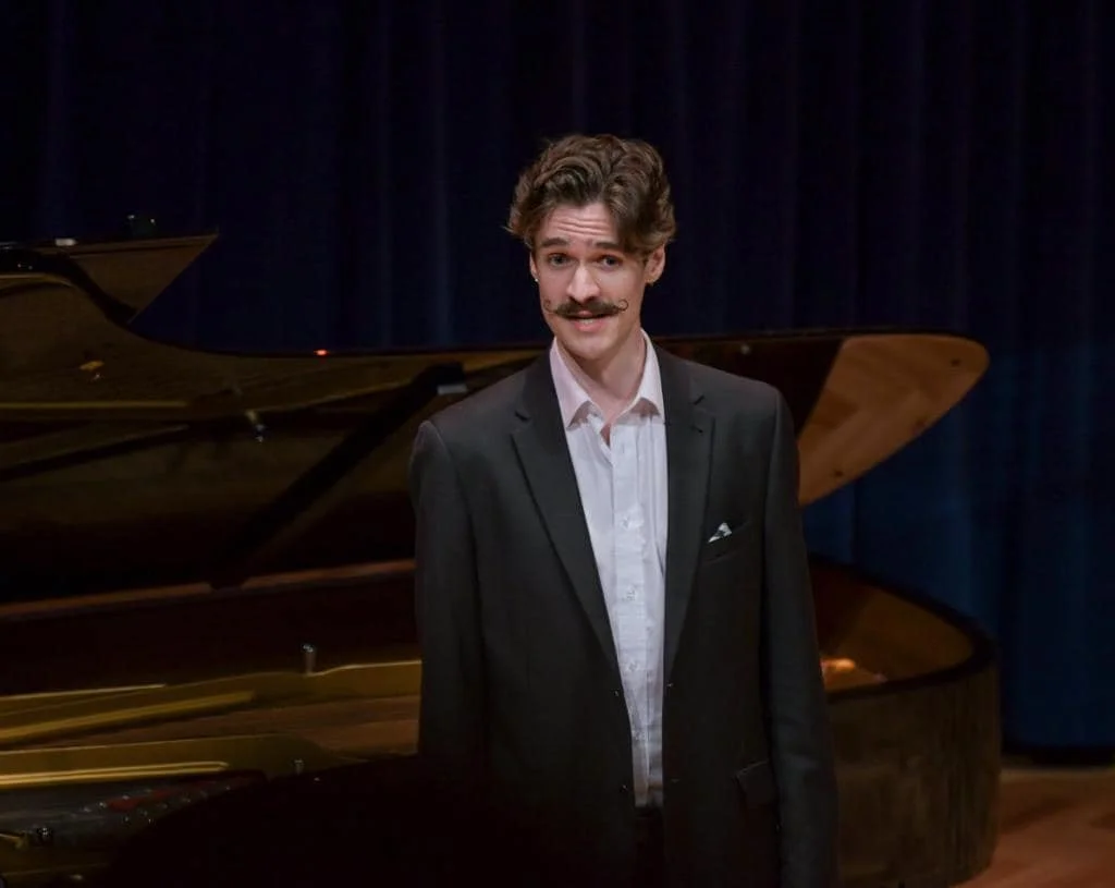 A man dressed in a black suit and white shirt standing in front of a grand piano on a stage with dark blue curtains.