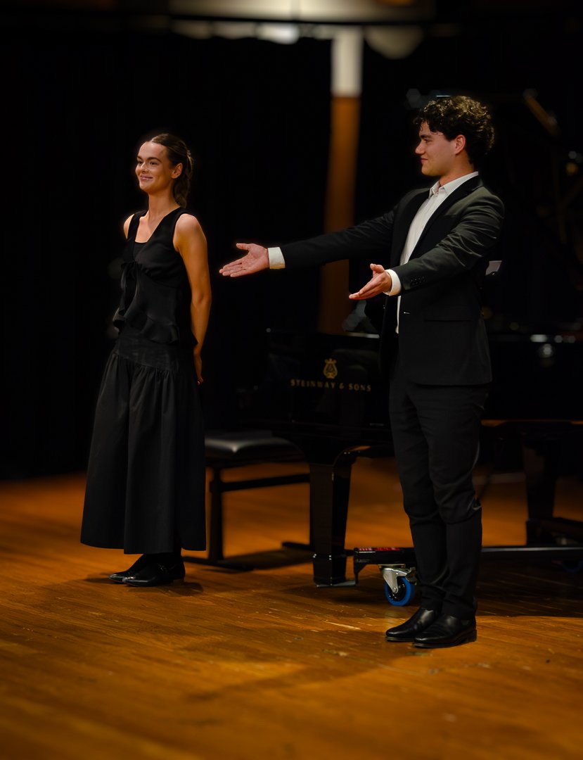A woman and a man in formal attire on a stage, with the man extending his hand towards the woman, next to a grand piano.