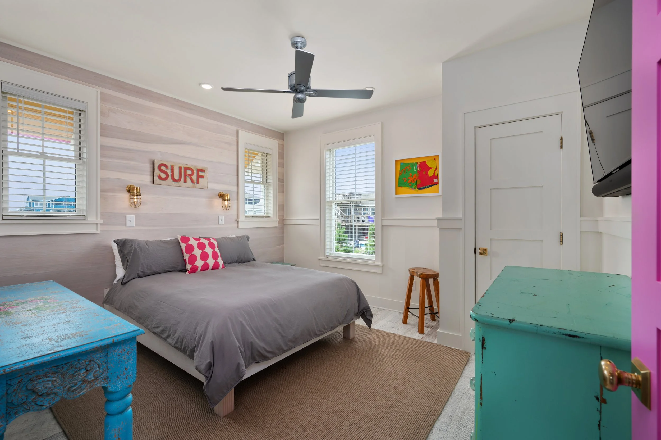 A bright bedroom with a bed covered in gray bedding, decorated with pink and gray pillows. There are three windows with white blinds, a chandelier fan, and artwork on the walls. The room includes a teal distressed dresser, a blue side table, and a wo