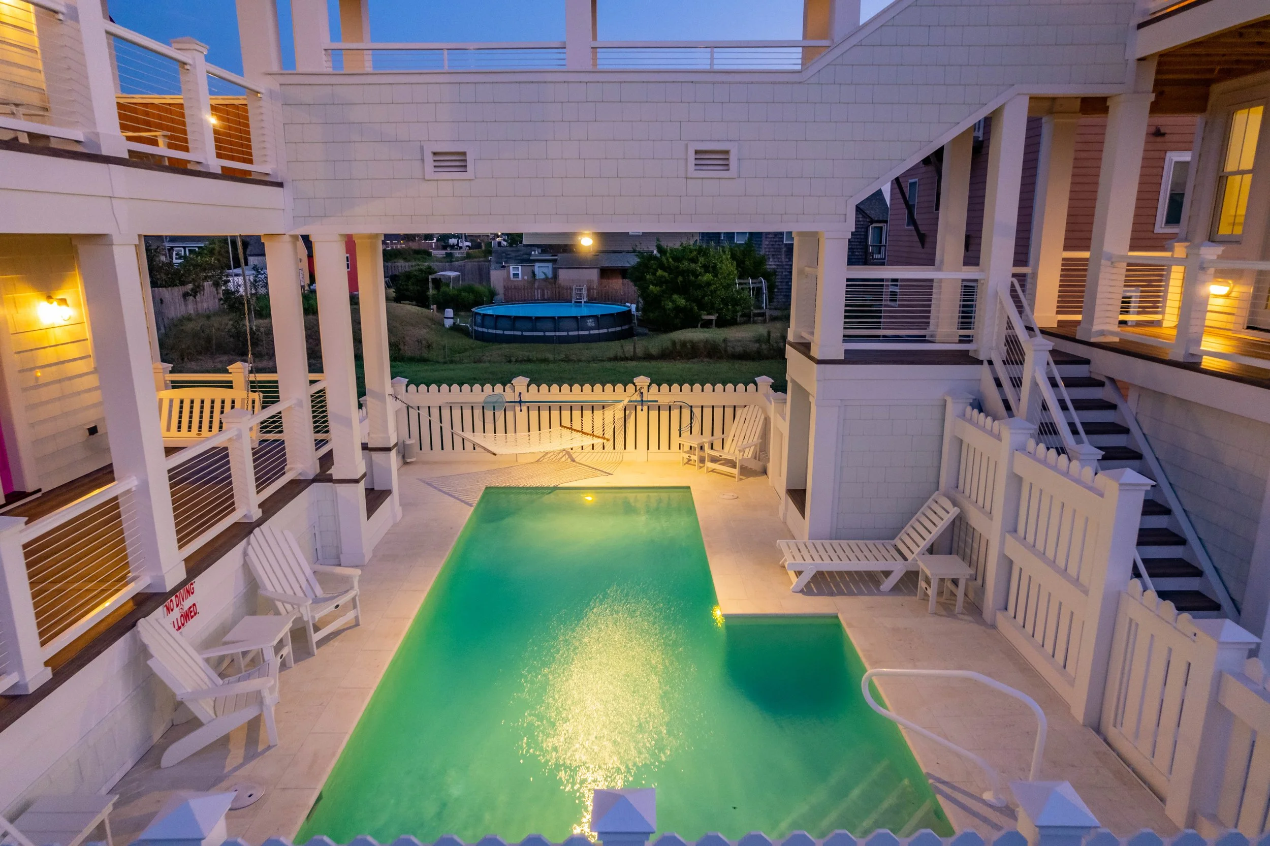An outdoor swimming pool illuminated with yellow lighting at night, surrounded by white Adirondack chairs, a hammock, and a fence with a backyard and a trampoline in the distance.