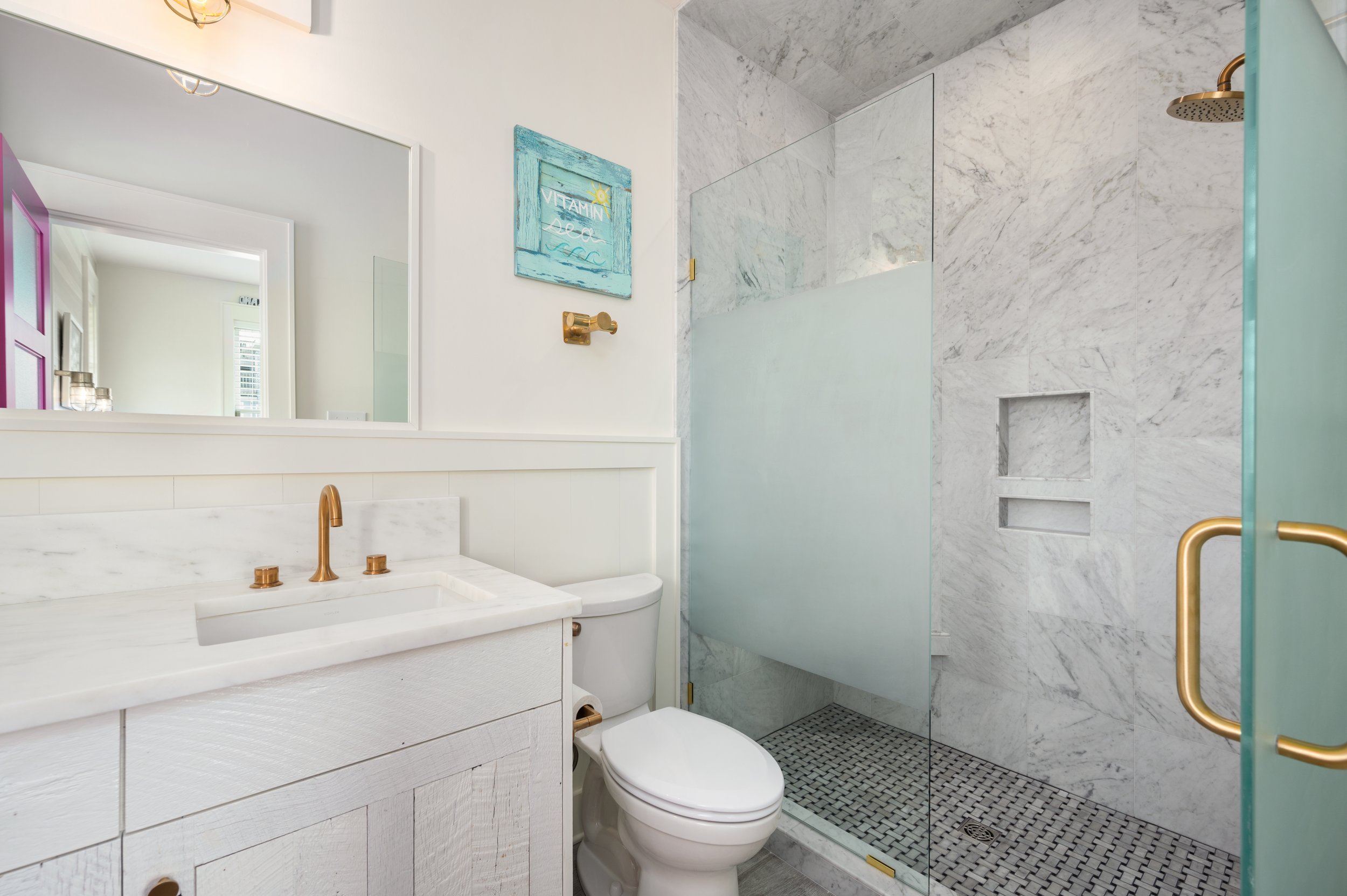 A modern bathroom with white walls and marble accents, featuring a white vanity with a marble sink, gold fixtures, a toilet, and a walk-in shower with marble walls and glass door.