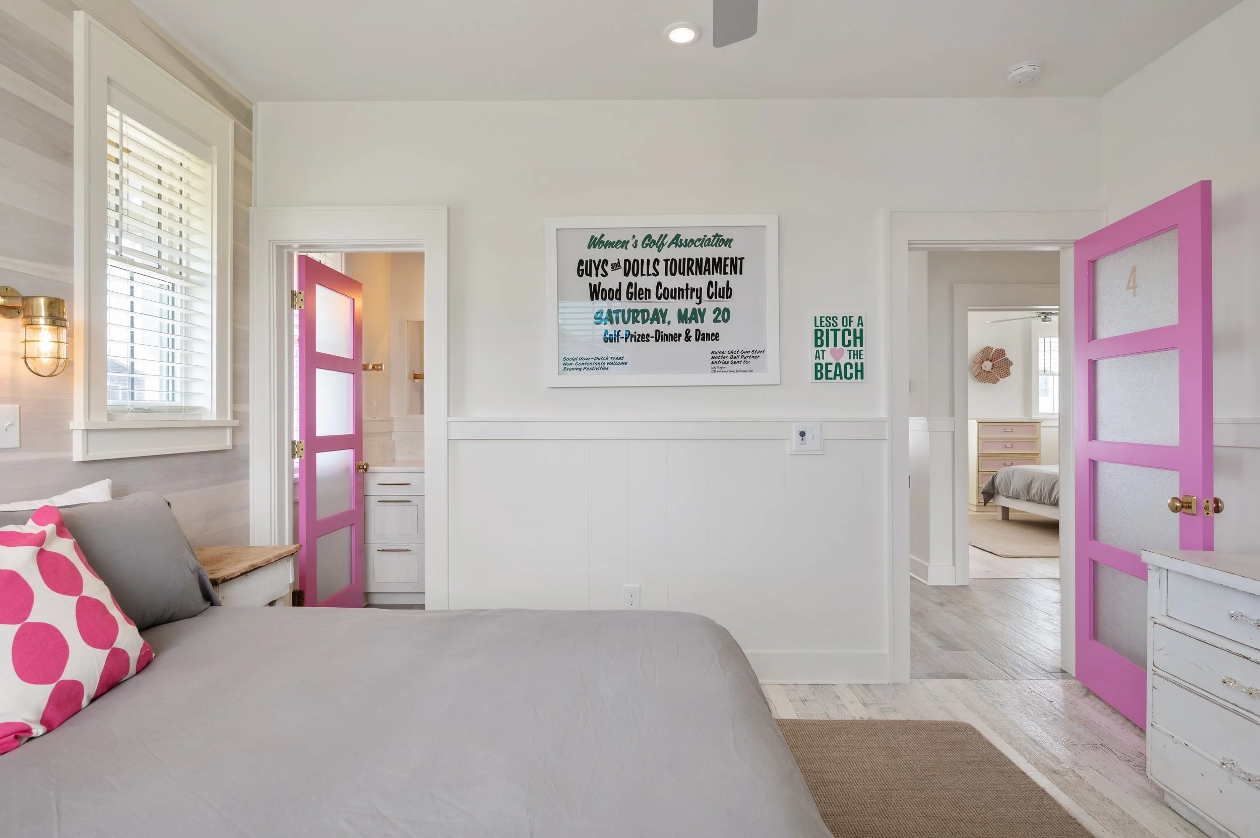 A bedroom with pink doors, a bed with pink and gray pillows, a window with white blinds, and framed signs on the wall, one advertising a golf tournament and another with humorous text.