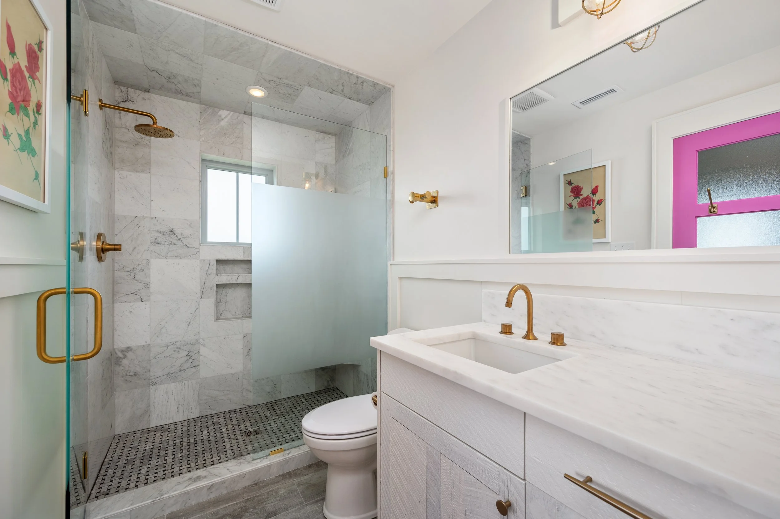 Modern bathroom with marble shower, white vanity with marble top, gold fixtures, and a large mirror.