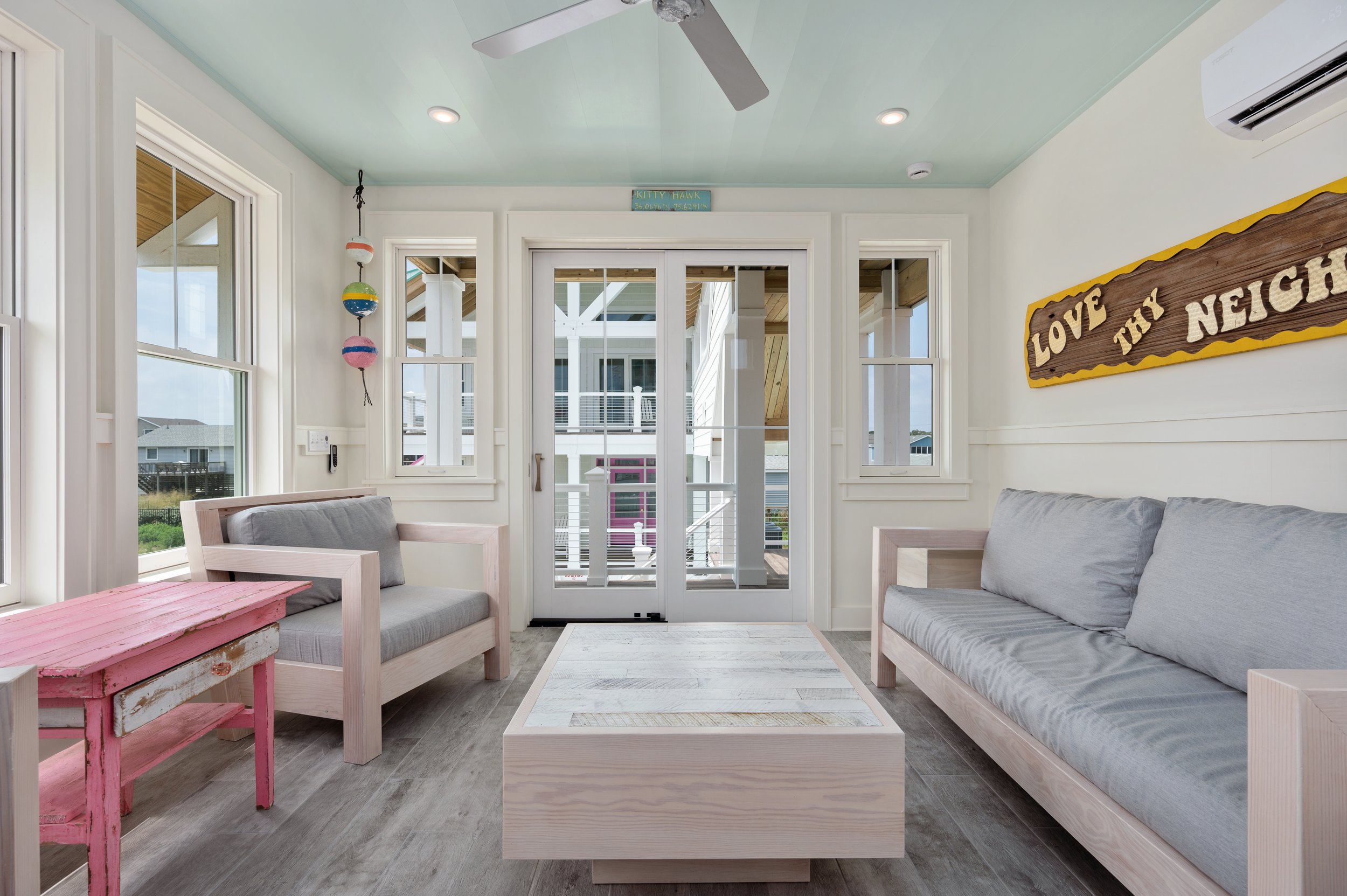 Bright living room with white walls and light wood furniture, including a large sofa, love seat, and a coffee table. Windows and glass doors lead to a balcony with outdoor furniture, and decor includes a sign that says 'Love thy neighbor' and a hangi