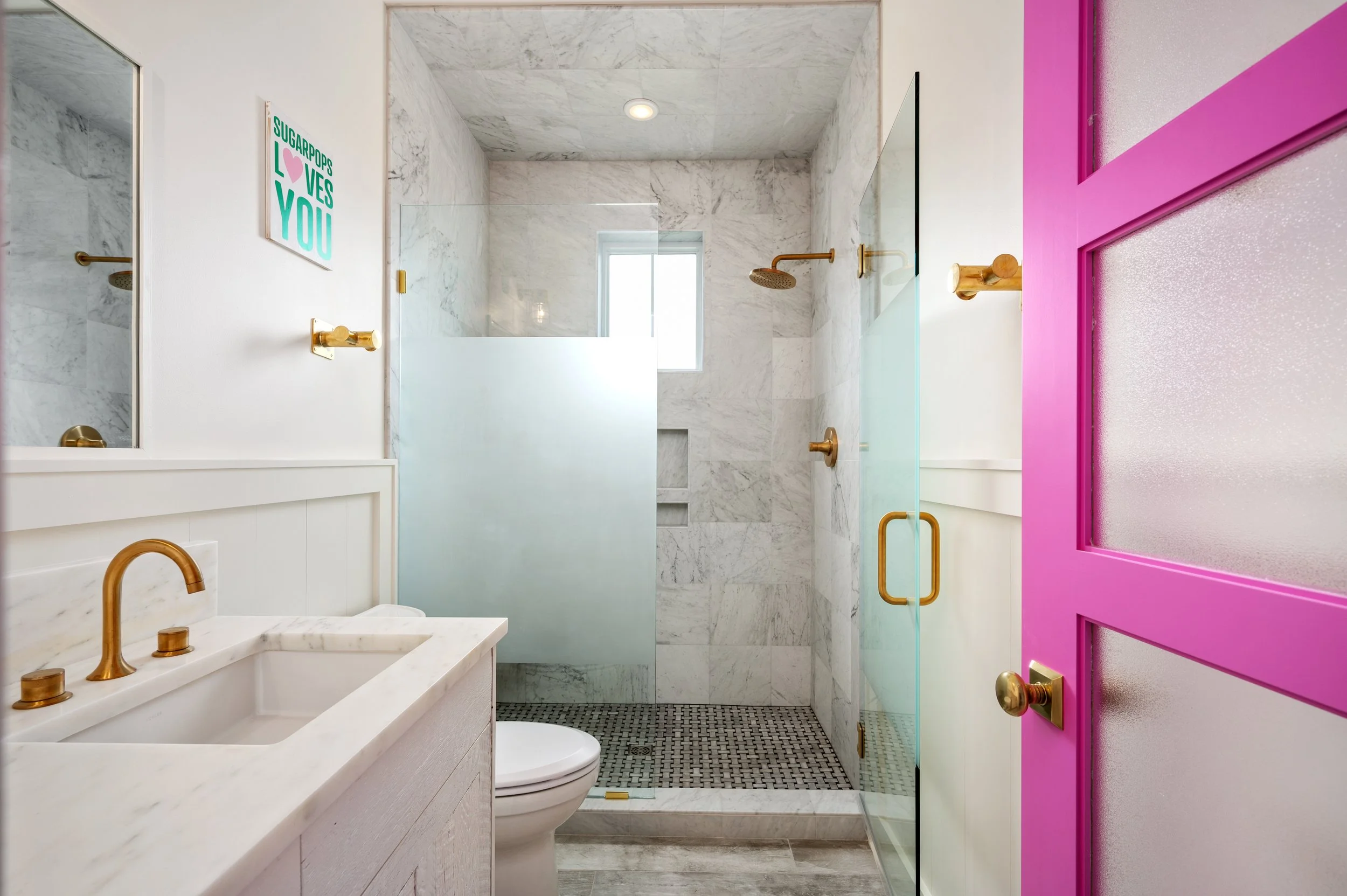 Modern bathroom with white marble shower, gold fixtures, pink door, white vanity, and a colorful sign that says "SUGARPOPS LOVES YOU."