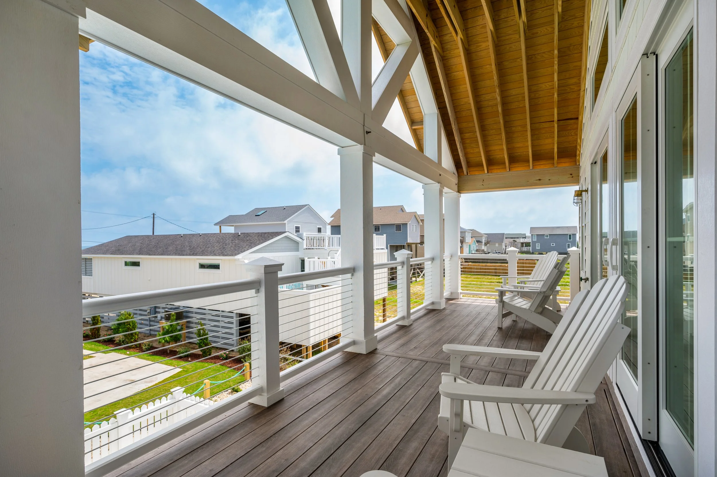 A spacious outdoor balcony with wooden flooring and white chairs, overlooking neighboring houses and a partly cloudy sky.