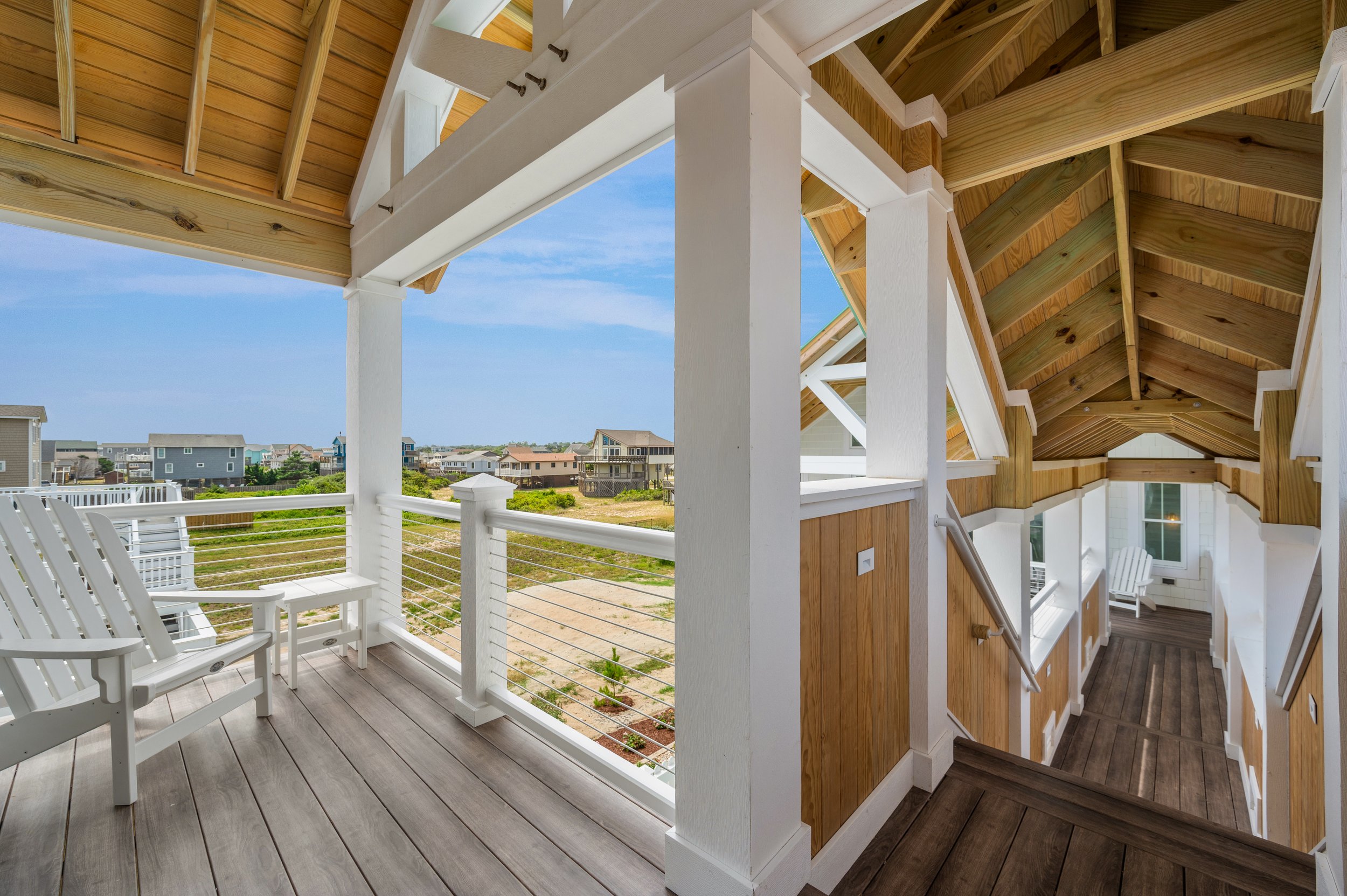 View from a house porch showing outdoor chairs and a railing, overlooking a grassy area with beach houses under a blue sky.