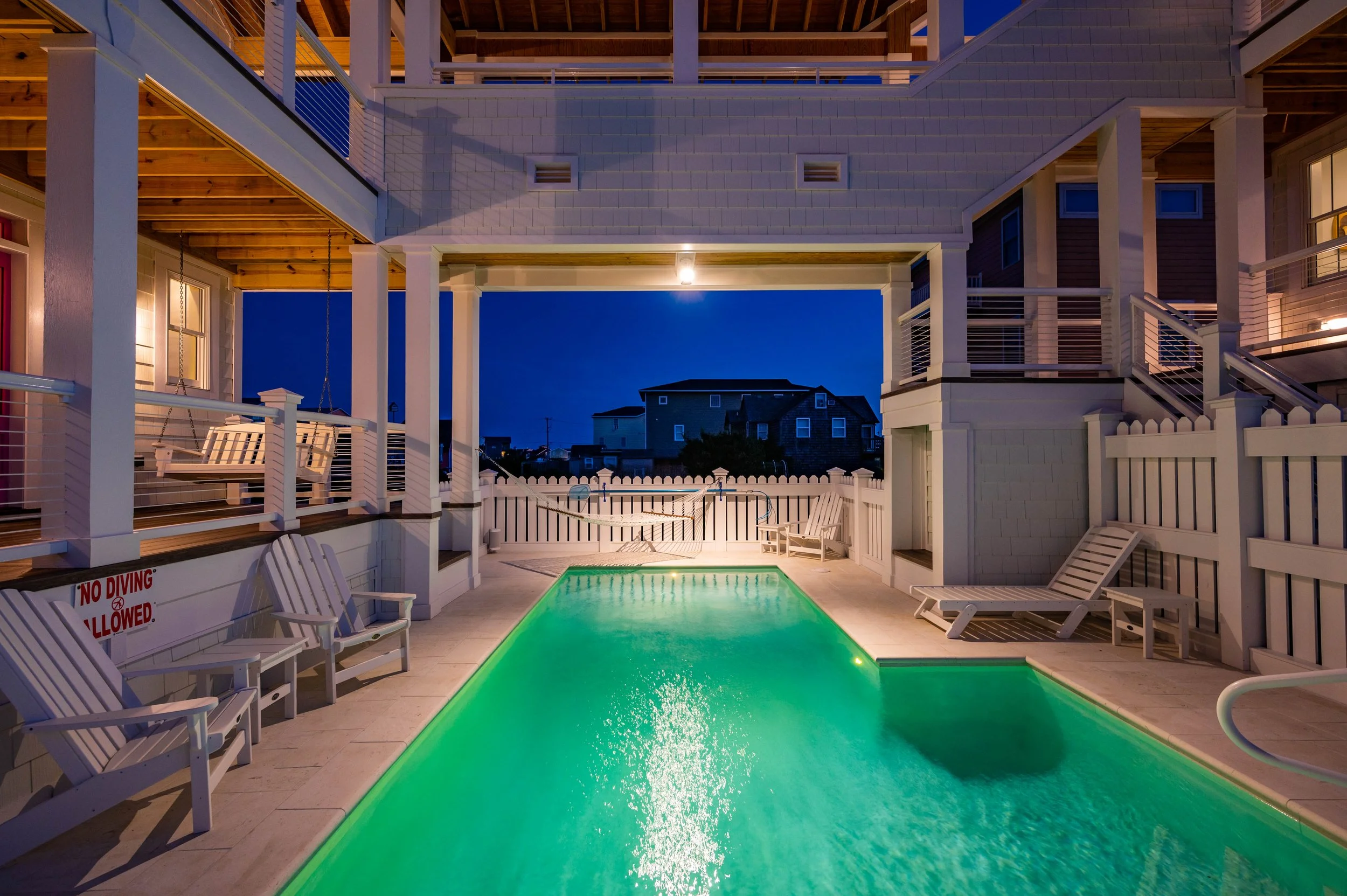 Night view of a small illuminated pool in a backyard with deck chairs, a swingset, and a white fence, with houses and the dark sky in the background.