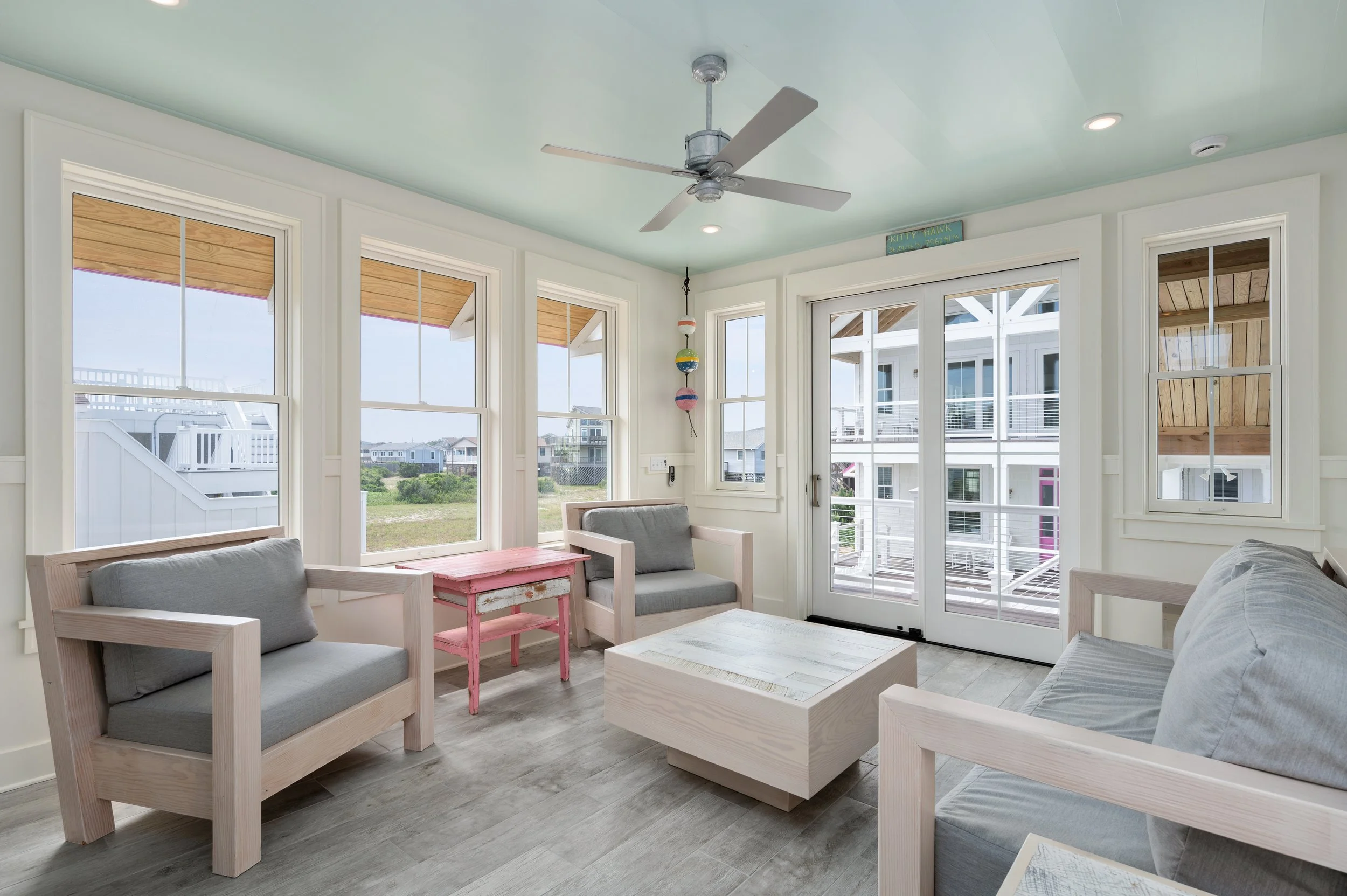 Bright sunroom with large windows, white furniture with gray cushions, a pink side table, and a ceiling fan.